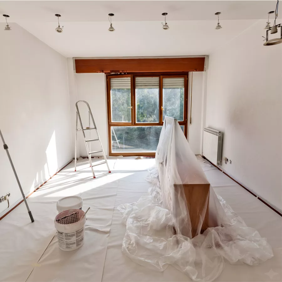 Room masked off before painting: floor fully sheeted, furniture wrapped in plastic, ladder, paint buckets and a roller tray ready to go