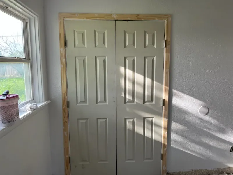 Double closet doors in new pine casing, prepped for painting, with a paint bucket on the windowsill